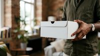 Man in olive green shirt holding white delivery box in modern loft workspace with brick walls and natural light, suitable for e-commerce, logistics, and small business concepts.
