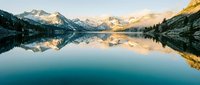 Serene mountain lake with snow-capped peaks reflecting in calm waters during golden hour sunrise, creating mirror symmetry in alpine wilderness landscape.