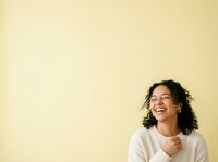 Young woman with curly dark hair and glasses, wearing a white long-sleeve shirt, smiling joyfully against a soft yellow background. Suitable for lifestyle, wellness, and professional content.