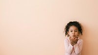 Young girl with curly dark hair and medium skin tone, wearing pink shirt, resting chin on hands against neutral beige background. Suitable for child development, education, and lifestyle content.