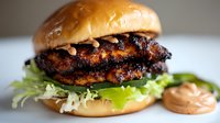 Grilled chicken sandwich with caramelized glaze on brioche bun, topped with shredded lettuce and special sauce, close-up view on light background.