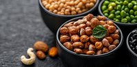 Assorted nuts and legumes in black ceramic bowls on dark stone background. Fresh hazelnuts, chickpeas, green peas and black beans for healthy protein snack.