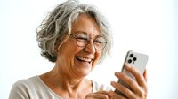 Senior woman with gray hair and glasses smiling while using smartphone for communication, technology, or social media on white background.