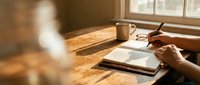 Young adult with medium skin tone writing in notebook at wooden desk by window with warm natural light, suitable for education, productivity, and lifestyle content.