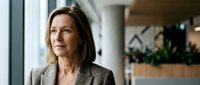 Mature woman with long brown hair and fair complexion wearing a gray blazer, standing in a modern office corridor with blurred background, suitable for corporate, business, and professional concepts.