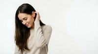 Young woman with long dark hair and South Asian ethnicity wearing a beige knit sweater, touching her hair with a gentle smile against a white background for portrait and lifestyle use.