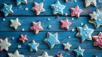 Star-shaped sugar cookies decorated with pink, blue and white royal icing on dark blue wooden background with small star sprinkles scattered around for festive holiday baking.