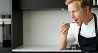 Man with reddish-blonde hair, Caucasian, wearing white shirt and black apron, tasting food with wooden spoon in modern kitchen setting.