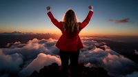 Young woman in red jacket celebrating success on mountain peak above clouds at dramatic sunset, arms raised in victory pose showing achievement and freedom.
