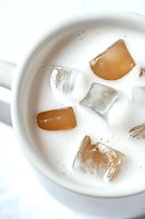 White ceramic mug filled with creamy milk and floating ice cubes, some clear and some with caramel-colored tint, creating refreshing summer drink composition.