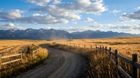 Rural dirt road with wooden fence leading through golden grassland toward mountain range under dramatic cloudy sky at sunset for travel and adventure themes.