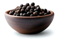 Black peppercorns in rustic brown ceramic bowl on white background, macro food photography showing texture and detail of aromatic spice ingredients.