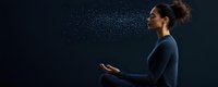Young mixed race woman in blue sweater meditating in lotus position while water droplets spray in air against dark background, side view wellness concept.