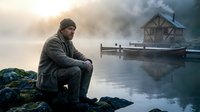 Contemplative man in warm clothing sitting by misty lake at dawn with wooden dock and boathouse. Peaceful morning solitude and reflection concept.