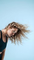 Young blonde woman with flowing wavy hair in motion against light blue sky background, expressing joy and freedom in casual summer outfit.