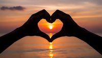 Hands forming heart shape silhouette against golden sunset over ocean water with romantic warm light reflection creating peaceful love symbol scene.