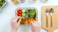 Hands in latex gloves preparing fresh vegetable meal prep container with tomatoes, cucumbers, greens and carrots. Food preparation and healthy eating concept.