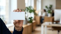Cropped view of male hand holding blank white card in modern office environment with blurred colleagues and plants in background for business messaging.
