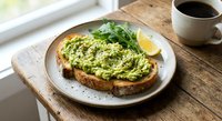Avocado toast on sourdough bread with herbs and lemon served on rustic plate with coffee cup on wooden table by window for healthy breakfast meal.