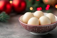Fresh white eggs in decorative ceramic bowl with red and gold Christmas lights and pine branches in soft focus background creating festive holiday mood.