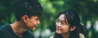 Young Asian couple sharing intimate moment outdoors with natural green bokeh background. Romantic connection between teenage boy and girl in casual clothing.