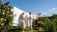 Young couple hanging white laundry on clothesline in sunny garden with blooming roses and vegetables, sustainable living and domestic chores concept.
