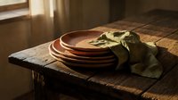 Stacked terracotta plates with green linen cloth on rustic wooden table in warm sunlight, suitable for food styling and tableware design.