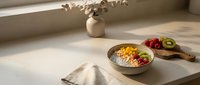 Overhead view of a nutritious breakfast bowl with granola, yogurt, and fresh berries on a minimalist white table with natural sunlight and dried flowers in a vase.