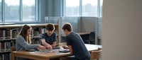 Three diverse students collaborating at library table with books and laptops, studying together in modern academic environment with natural window lighting.