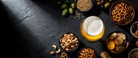 Beer glass with foam surrounded by various snacks including pretzels, nuts, and appetizers on dark slate background for oktoberfest celebration.