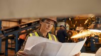 Male engineer in safety glasses and yellow reflective vest reviewing blueprints at industrial manufacturing facility with machinery and workers in background.
