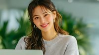 Young Asian woman smiling warmly in cozy gray sweater at home with green plants in background for lifestyle and wellness content.