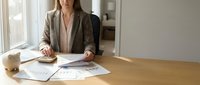 Professional businesswoman in beige blazer working with documents and calculator at modern office desk, reviewing financial reports and paperwork.