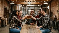 Young diverse friends toasting with craft beer glasses in rustic brewery taproom with exposed brick walls and industrial lighting for social gatherings.