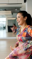 Young Southeast Asian woman in colorful patterned wrap dress smiling in modern office lobby with colleagues in background, suitable for corporate, diversity, and workplace culture content.