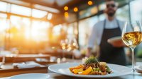 Elegant plated dish with fresh garnish and white wine glass in upscale restaurant setting, warm ambient lighting with blurred professional server in background.