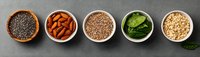 Healthy superfood ingredients in ceramic bowls on grey concrete background - chia seeds, almonds, quinoa, spinach and oats arranged in row.