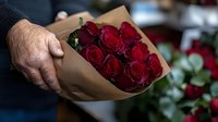 Male hand holding elegant bouquet of fresh dark red roses wrapped in brown kraft paper, romantic gift concept captured in moody lighting.