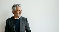 Mature man with gray hair and facial hair wearing dark blazer and navy sweater, looking away thoughtfully against white background. Suitable for business, corporate, and lifestyle design projects.