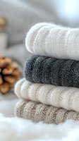 Stack of cozy knitted sweaters in neutral tones with cable pattern texture, folded neatly on soft white surface creating warm autumn winter fashion concept.