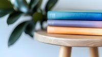 Colorful stack of notebooks on wooden stool against blurred green plant background, minimalist home office or study corner setup for planning and organization.