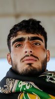 Close-up portrait of young man with dark beard and defined eyebrows wearing patterned scarf, looking up with confident expression against light background.