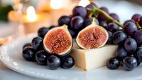 Fresh ripe figs cut in half revealing pink flesh alongside dark purple grapes and cheese on white plate with soft candlelight bokeh background.