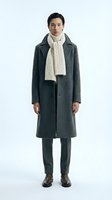 Young East Asian man wearing charcoal wool coat with cream cable-knit scarf and dark trousers, standing against white background for fashion retail and apparel marketing.