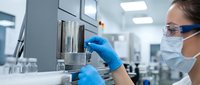 Young female scientist or laboratory technician wearing protective eyewear, face mask, and blue latex gloves conducting research or analysis in a modern sterile lab environment.