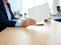Middle-aged man in dark business suit holding blank white card at wooden desk in modern office environment with colleagues working in background.
