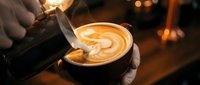 Professional barista creating latte art in coffee cup with steamed milk, warm cafe atmosphere with bokeh lights in background for hospitality concepts.