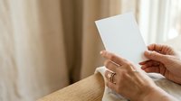 Mature woman with light skin tone holding blank white card in hands at wooden table with beige curtains in background for greeting, invitation, or announcement mockup.