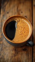 Fresh brewed coffee with golden crema foam in dark ceramic cup on rustic wooden table, top view close-up shot showing rich texture and bubbles.