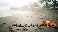 Aloha written in sand on a tropical beach with colorful hibiscus flowers, palm trees, and ocean waves in the background.